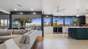 A modern living room with a large sofa, a kitchen area with a bar, and a view of palm trees. - Cactus & Sky Escape with POOL and SPA! (Tucson)