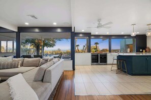 Ein modernes Wohnzimmer mit einer großen Couch, einer Küchenzeile mit Bar und Blick auf Palmen.