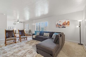 Ein Wohnzimmer mit einem Ecksofa, zwei Sesseln, einem Couchtisch, einer Stehlampe und Bildern an der Wand.
