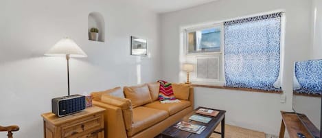 A living room with a sofa, a coffee table, a lamp, a radio, a TV, and a window with patterned curtains.