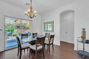 Uma área de refeições com mesa e cadeiras de vidro, um candelabro e vista para um jardim exterior através de portas de vidro.