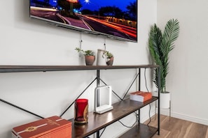Een flatscreen televisie aan een wand bevestigd, een houten plank met boeken en een rode vaas, en een potplant.