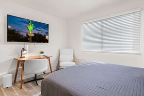 Een slaapkamer met een bed, bureau, stoel, televisie aan de muur, en een raam met zonwering.