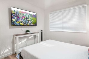Ein Schlafzimmer mit einem Bett, einem an der Wand befestigten Fernseher, einem Fenster mit Jalousien und einem kleinen Tisch mit einer Pflanze.