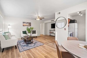 Ein modernes Wohnzimmer mit Deckenventilator, eine Essecke mit rundem Tisch und eine Küche mit Kühlschrank.