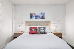 Um quarto com parede de tijolos brancos, uma cama grande com duas almofadas, duas mesas de cabeceira com abajures e três quadros abstratos acima da cama.