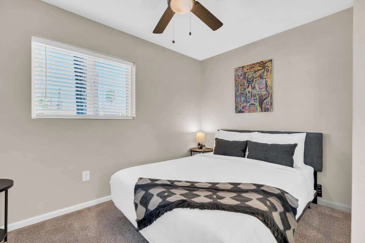 Une chambre à coucher avec un lit, un ventilateur de plafond, une fenêtre avec des stores, une table de chevet avec une lampe et un tableau au mur.