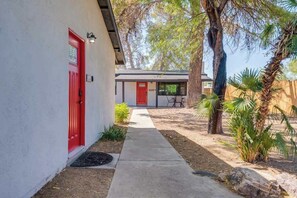 Un camino que lleva a un edificio con puerta roja, un pequeño espacio al aire libre para sentarse y una palmera.