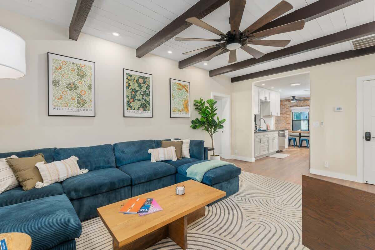 A living room with a blue sectional sofa, a wooden coffee table, and a ceiling fan. There are framed botanical prints on the wall and a potted plant.