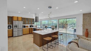 A modern kitchen with a central island, bar stools, and a large window overlooking a garden. - Desert Vibes with Pool, Sauna & Game Room! (Tucson)