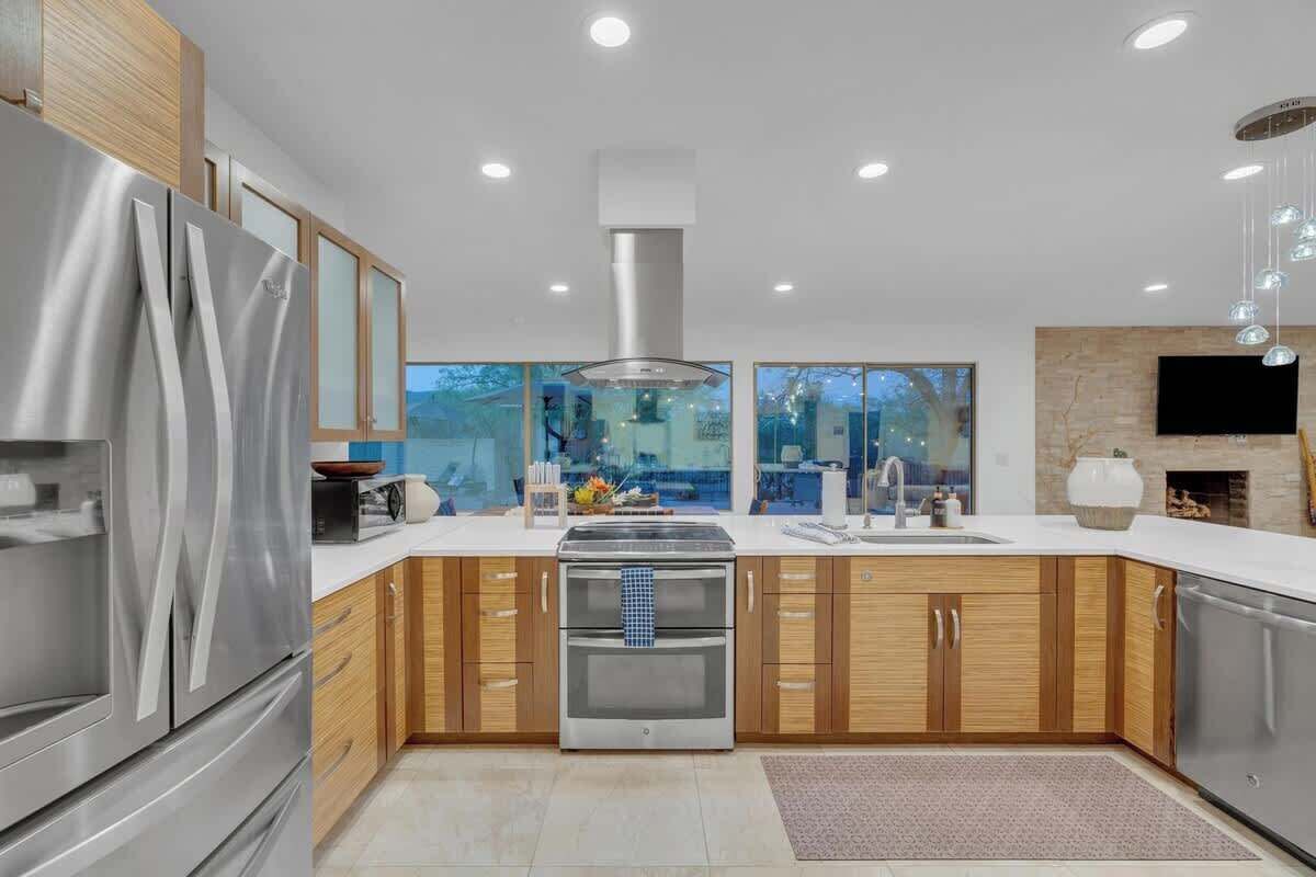 A modern kitchen with stainless steel appliances, wooden cabinets, a large window, and a flat-screen TV.