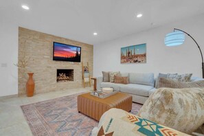 A living room with a stone wall, a flat-screen TV, a sofa, a patterned rug, and a modern floor lamp. - Desert Vibes with Pool, Sauna & Game Room! (Tucson)