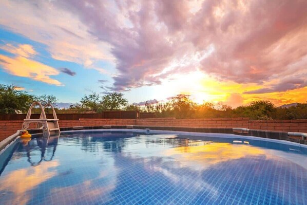 En swimmingpool med en stige, omgivet af en murstensmur, og en solnedgang i baggrunden.
