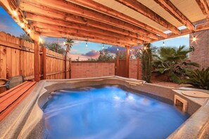 Ein Whirlpool unter einer Holzpergola mit Lichterketten, umgeben von einem Holzzaun und Palmen.