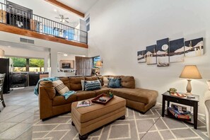 A spacious living area with a sectional sofa, ottoman, and a coffee table. There is a balcony with a railing, a kitchen in the background, and artwork on the walls. - Modern Loft with Pool & Hot Tub-Views! (Tucson)