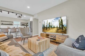 A modern living room with a large flat-screen TV, a sofa, a coffee table, and a dining area with chairs and a kitchen in the background. - The Hidden Gem-5 mins to U of A with Hot Tub (Tucson)
