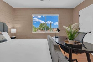 Une chambre à coucher comprenant un lit, un bureau, une lampe, une plante en pot et une fenêtre donnant sur des immeubles et des arbres.