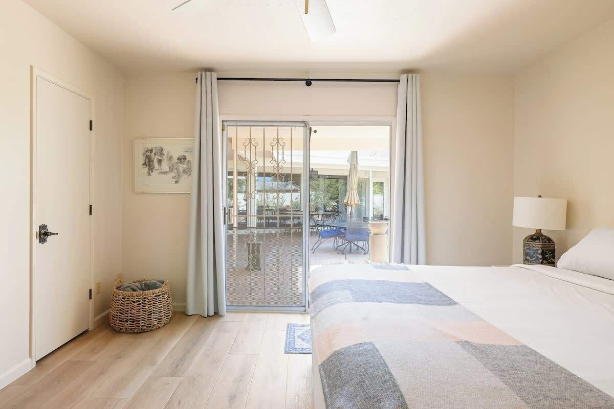 A hotel room with a bed, a door, a wicker basket, a sliding glass door to an outdoor area, and a framed picture on the wall.