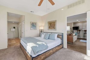 Ein Schlafzimmer mit einem Bett, einem Deckenventilator, einem Gemälde und Blick zur Küche.