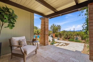 A covered patio area with a brick pillar, a cushioned chair, and a view of a pool and desert landscape. - Catalina Foothills Luxe Retreat with Heated Pool! (Tucson)