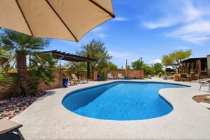 A swimming pool with a curved edge, surrounded by a concrete deck, palm trees, and a pergola with a beige canopy. - Catalina Foothills Luxe Retreat with Heated Pool! (Tucson)