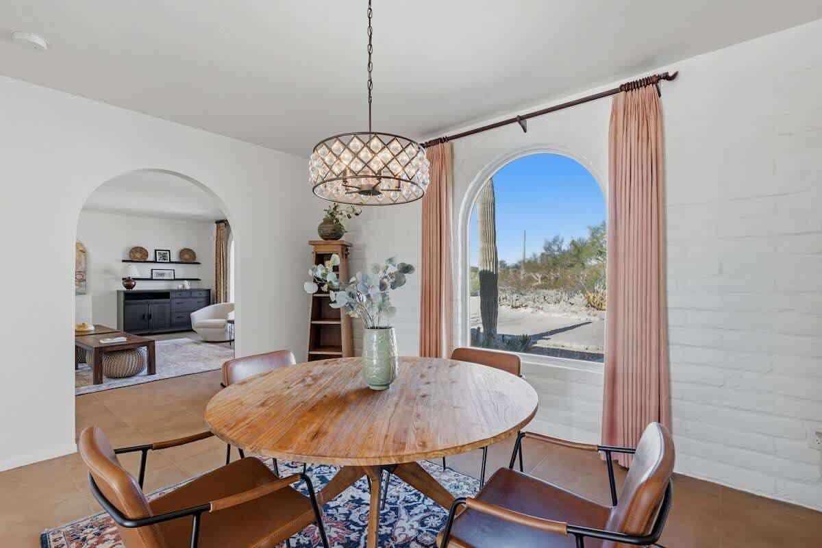 A dining area with a round wooden table, six chairs, a hanging light fixture, and a view through a large window.