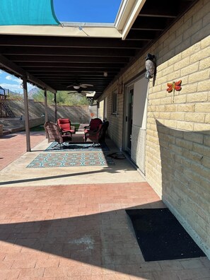 Ein überdachter Patio-Bereich mit Gartenmöbeln, einem gemusterten Teppich und einem gepflasterten Weg, der zu einer Garage führt.
