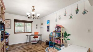 Huoneessa on puulattia, valkoinen pöytä, ikkuna, kattotuuletin ja kirjahylly.