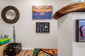 Un pasillo con un espejo de madera, una pared con un cuadro de colores, un letrero enmarcado que enumera animales, un caja de almacenamiento verde, un baúl negro y una alfombra colorida.