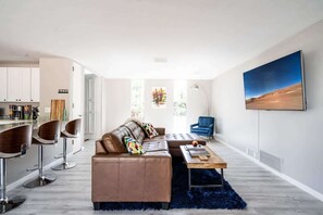 Ein modernes Wohnzimmer mit Sofa, Couchtisch und Barhocker. An der Wand hängt ein Gemälde und im Hintergrund ist eine Küchenzeile zu sehen.