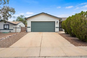 A single-story house with a green garage door, a driveway, and a small tree to the right. - Cozy & Pet Friendly & Peloton! (Tucson)
