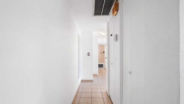 A narrow hallway with white walls and a tiled floor, leading to a room with a visible door and a vent on the ceiling. - Sunny & Bright Charm with Community Pool (Tucson)