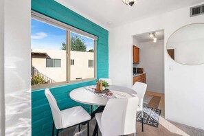 A room with a round table, white chairs, a window with a teal accent wall, and a kitchen area visible through an open door. - Sunny & Bright Charm with Community Pool (Tucson)