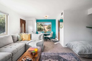 A modern living room with a grey sofa, a coffee table with books and a plant, a dining area with a teal accent wall, and a large window. - Sunny & Bright Charm with Community Pool (Tucson)
