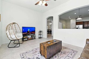 A living room with a television, a wooden coffee table, a hanging chair, and a kitchen area in the background. - Desert Solace Retreat w/Pool/Spa (Tucson)