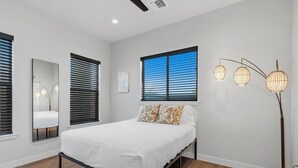 Ein Schlafzimmer mit einem Bett, einem Fenster mit Jalousien, einer stehenden Stehlampe und einem Spiegel.