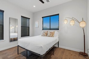A bedroom with a bed, a window with blinds, a standing floor lamp, and a mirror. - Modern Tucson Home Near UA & Downtown! (Tucson)