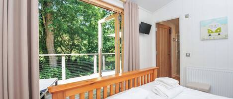 A bedroom with a large window, wooden headboard, a bed with white linens, a TV mounted on the wall, and a door leading to a shower area.