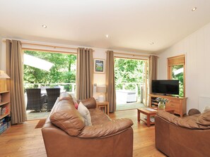 A spacious living room with a leather sofa, a television, and a view of greenery through large windows.