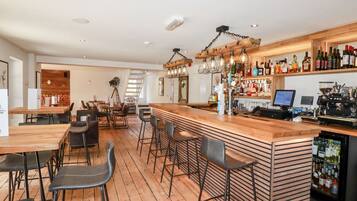 Zona de bar con suelo de madera, taburetes, una barra con una máquina de café y estantes con botellas.