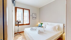 Una habitación de hotel con una cama, una mesita de noche con lámpara, una ventana con cortinas, un ventilador de pie y un cuadro colgado en la pared.