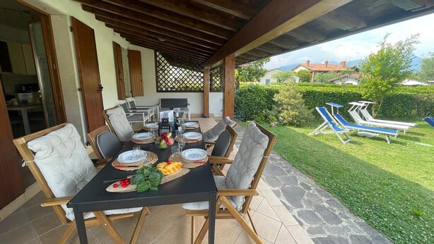 Un patio al aire libre con mesa lista para comer, rodeado de sillas y vistas a un jardín con sillones de descanso.