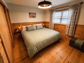 A bedroom with a wooden headboard, a bed with a green bedspread, a nightstand with a lamp, a window with patterned curtains, and a wall-mounted picture.
