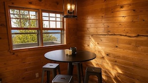 Interior de madera con una mesa redonda, dos taburetes, una ventana y una lámpara colgante.