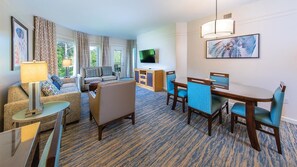 Habitación de hotel con sala de estar, una mesa de comedor con sillas, un televisor y un cuadro en la pared.
