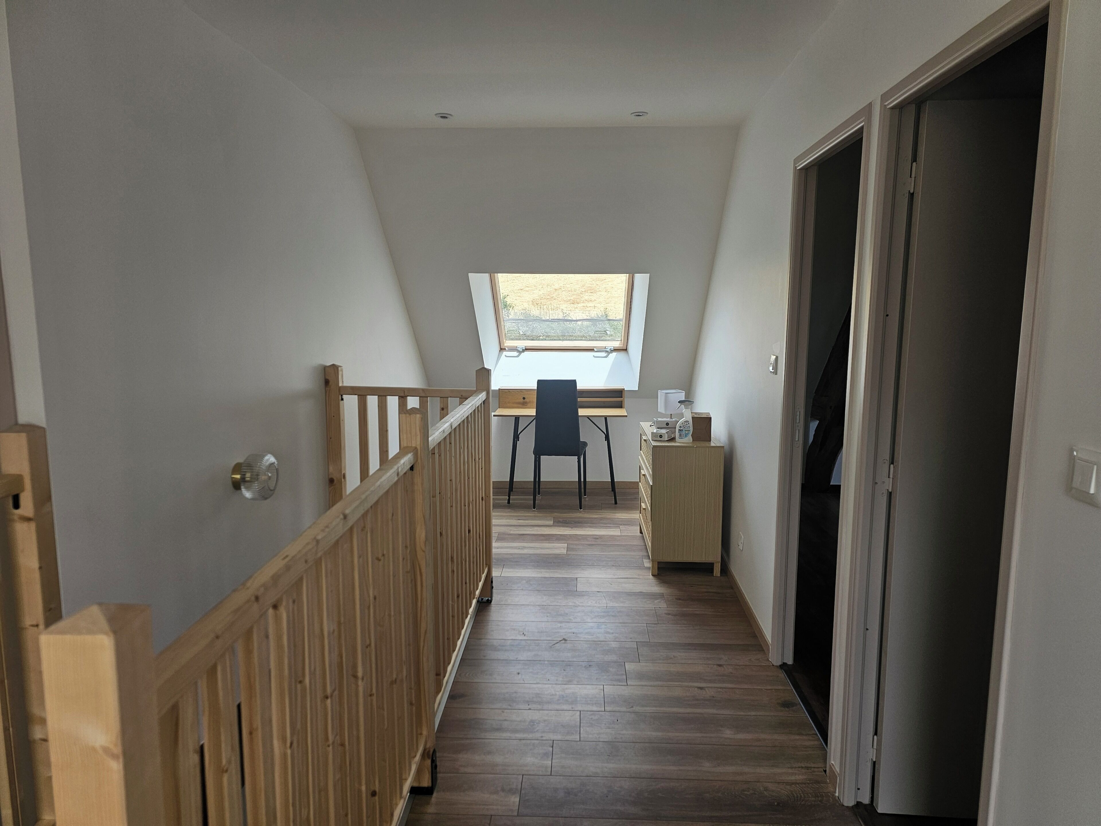 A narrow hallway with a wooden railing, a desk with a chair, and a window with a view of the outdoors.