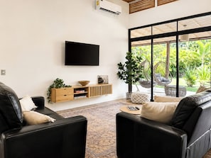 A modern living room with a flat-screen TV, a wooden console table, and a large window with a view of greenery. - 3 Bed, 2 Bath, AC, Family& Pet-Friendly Modern Jungle Oasis w/Private Pool (Puerto Viejo de Telemanca)