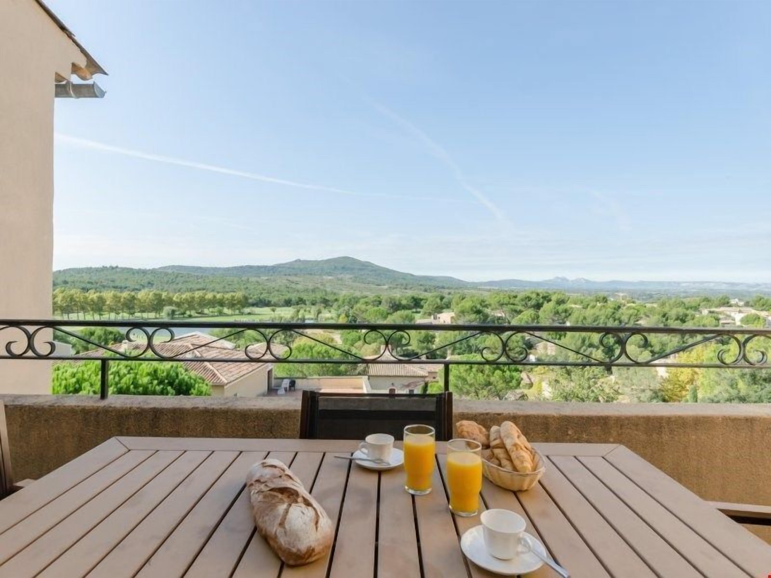 Un balcon avec une table préparée pour le petit-déjeuner, un pain, un verre de jus d’orange et un bol de viennoiseries.