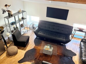 A modern living room with a black leather sofa, a glass coffee table, a round ottoman, a black armchair, a wooden floor, a metal shelving unit, and a flat-screen TV mounted on the wall. - L'Antonin - Clim mobile - Metro A et D (Lyon)