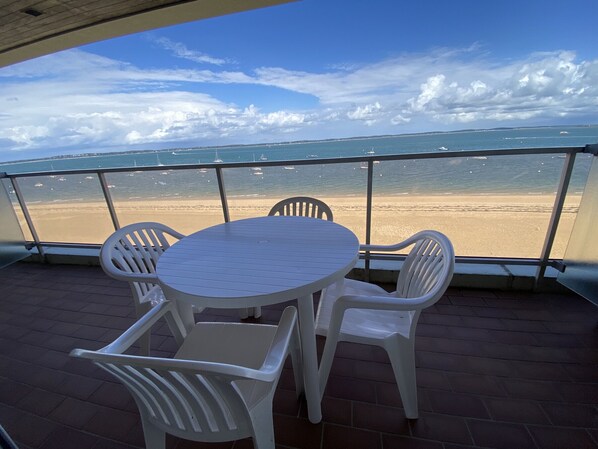 Een balkon met een ronde tafel en vier stoelen, met uitzicht op een strand met boten en een heldere lucht.